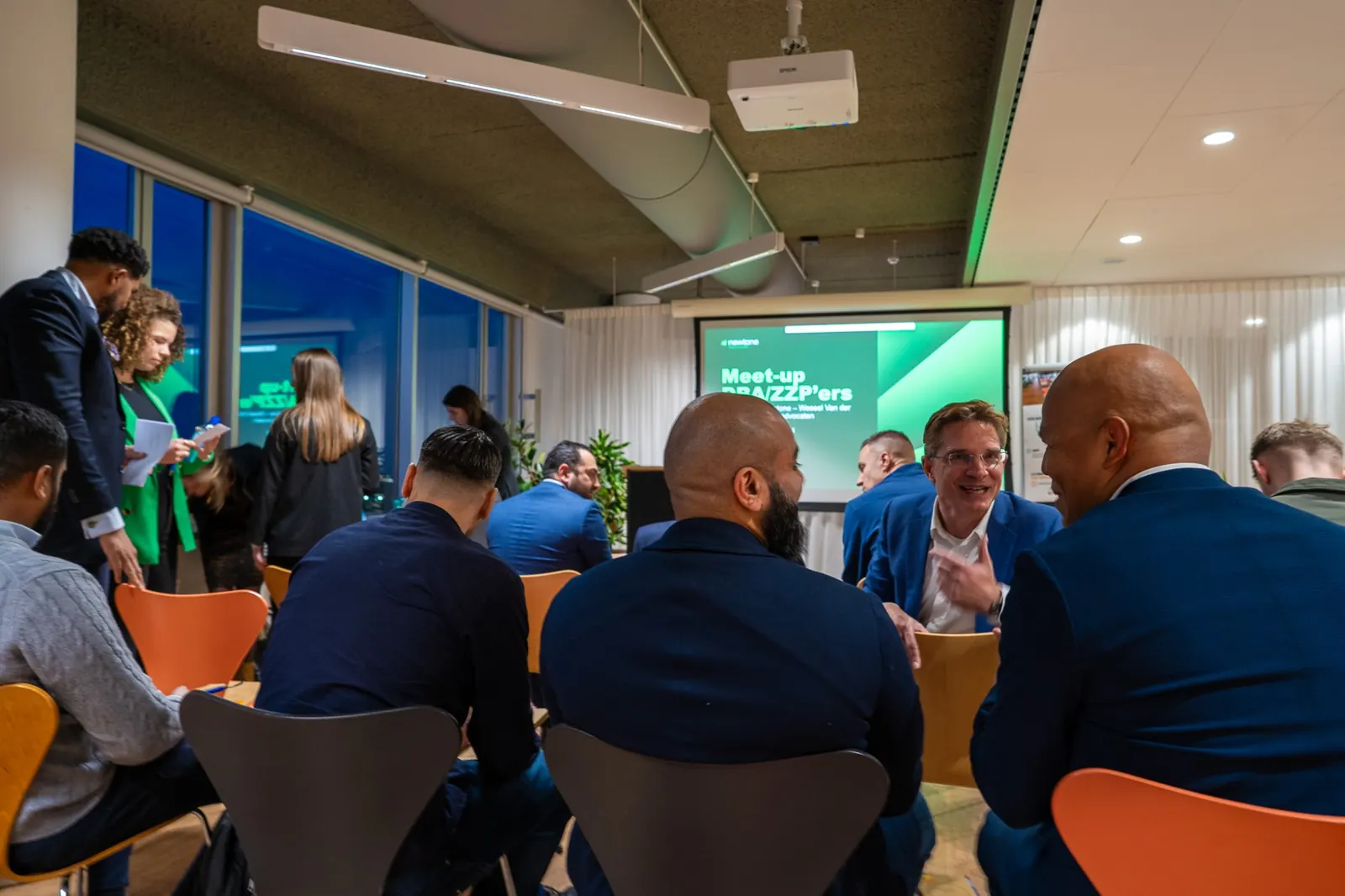 Deelnemers in gesprek met elkaar in de zaal, met het presentatiescherm op de achtergrond
