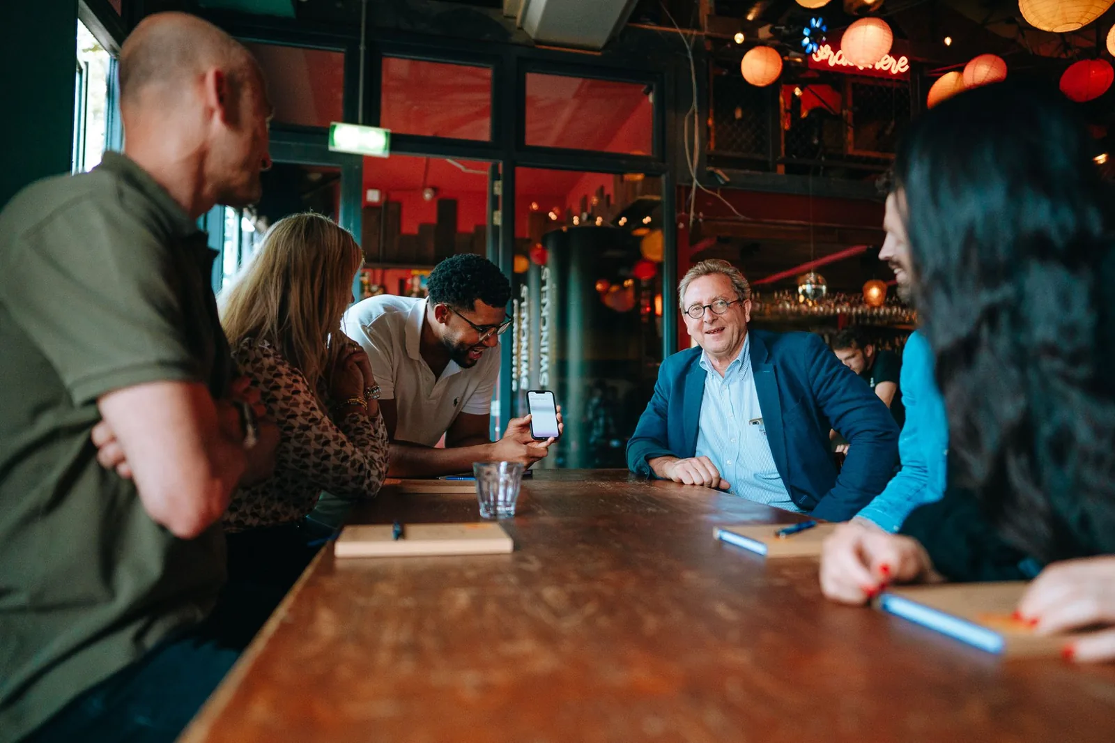 Deelnemers lachend in gesprek aan een lange houten tafel, een van hen laat iets zien op zijn telefoon